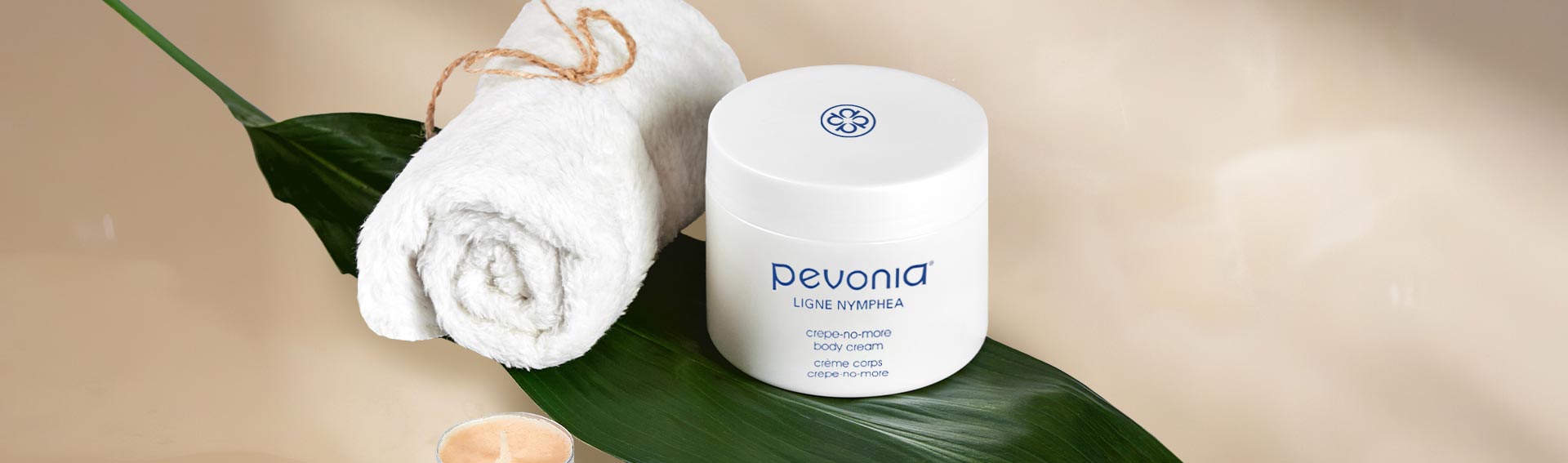 Pevonia's Crepe-No-More Body Cream on top of a leaf, next to a rolled spa towel and candles.